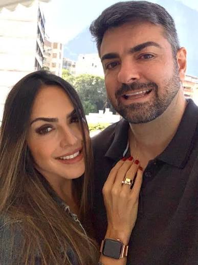 Um selfie em close-up do casal Luana e Leandro Piquet, sorrindo e olhando para a câmera. Suely está à esquerda, com longos cabelos castanhos e uma jaqueta jeans, apoiando a mão esquerda sobre o peito de Leandro, revelando um anel de casamento de ouro e um relógio inteligente dourado. Leandro está à direita, com barba e cabelos grisalhos curtos, vestindo uma camisa polo cinza escura. Eles estão em uma área externa urbana, com edifícios e árvores ao fundo sob um céu claro.