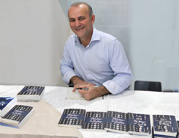 Fotografia em cores de Hélder Salomão sentado em uma mesa coberta por uma toalha branca, sorrindo e segurando uma caneta enquanto autografa um livro. Na mesa e ao seu lado, há vários exemplares organizados de suas obras publicadas.
