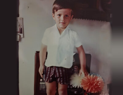 Fotografia em cores antigas de Hélder Salomão ainda criança, em pé, vestindo camisa social branca de manga curta e bermuda escura estampada, com elementos decorativos ao fundo.