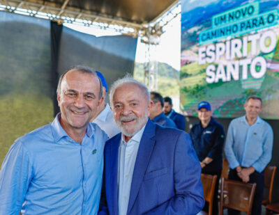 Fotografia em cores de Hélder Salomão, vestindo camisa social azul claro, e Luiz Inácio Lula da Silva, vestindo paletó azul marinho, sorrindo lado a lado. Ao fundo, vê-se um palco e um banner com a inscrição 'UM NOVO CAMINHO PARA O ESPÍRITO SANTO', indicando um ato público ou de campanha.