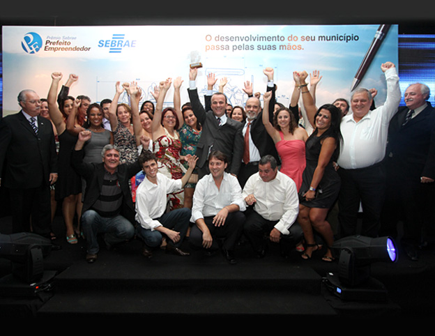 Fotografia em cores de um grupo grande de pessoas em um palco durante uma cerimônia de premiação, com participantes erguendo os braços e troféus em sinal de comemoração.