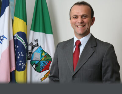 Fotografia em meio corpo de Hélder Salomão sorrindo, vestindo terno cinza, camisa social branca e gravata vermelha, posado à frente de bandeiras institucionais.