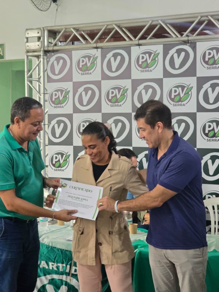 Uma fotografia colorida em ambiente interno mostra três pessoas em pé. Ao centro, uma mulher recebe um certificado de dois homens. Atrás deles, há um painel quadriculado com a logomarca repetida