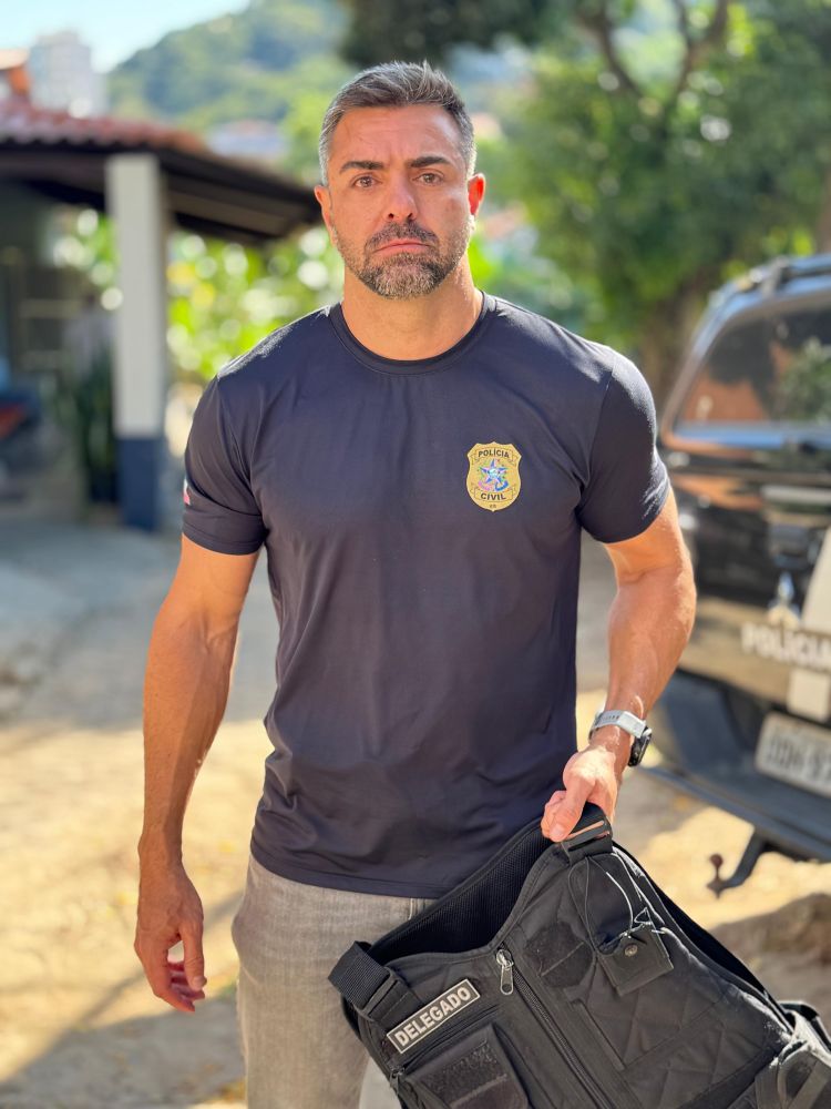 Fotografia de corpo inteiro do Delegado Leandro Piquet em pé, ao ar livre. Ele veste uma camiseta preta de manga curta com o brasão da Polícia Civil do ES no peito e calça cinza, segurando um colete tático preto com a identificação "DELEGADO" na altura da mão. Ao fundo, aparece parcialmente uma viatura policial caracterizada.